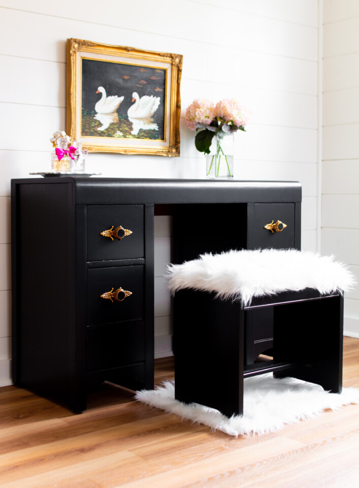 Angled view of staged coal black desk with flowers and pink perfume bottles on top, faux fur covered stool with gold accents, painting of swan hanging above dresser.