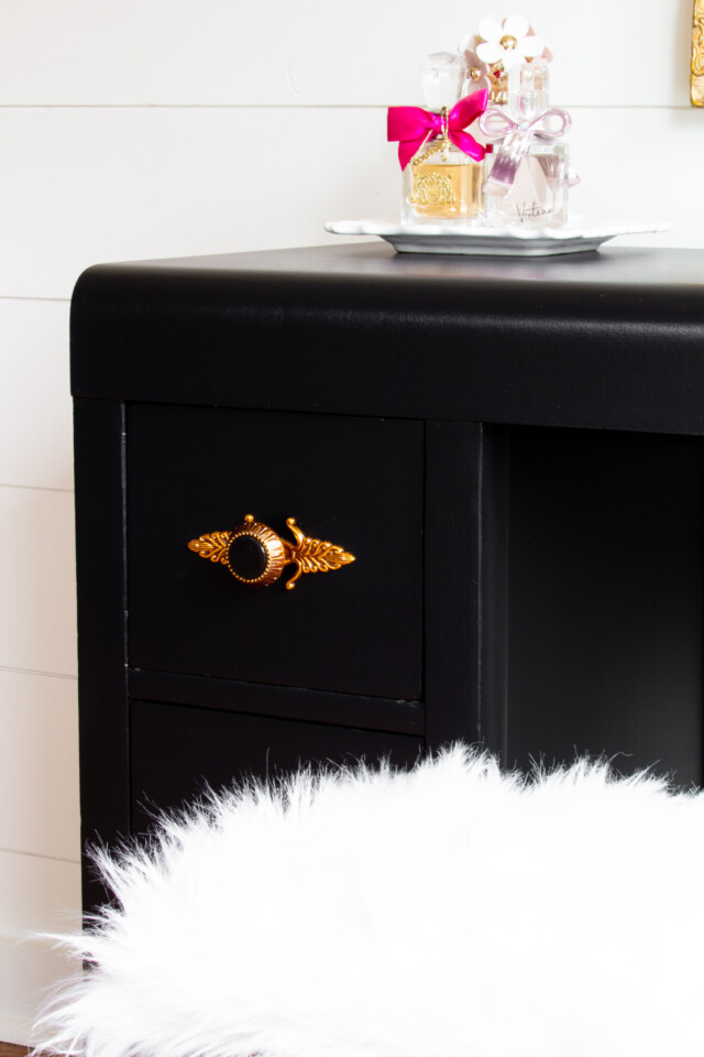 Close up image of deco desk painted in coal black, staged with pink perfume bottles on top and a faux fur stool