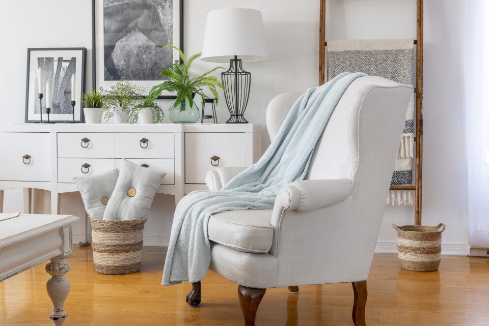 Side view photo of completed chair staged with a light blue blanket, basket of pillows, behind is a white dresser with plants and a blanket ladder.