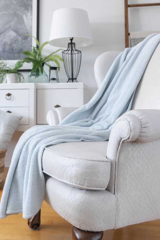 Close up side view of painted chair, staged with a light blue blanket, behind is a white dresser with plants