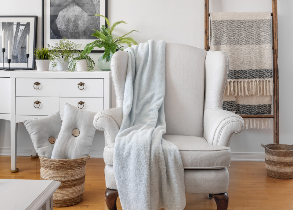 Front facing photo of completed chair staged with a light blue blanket, basket of pillows, behind is a white dresser with plants and a blanket ladder.