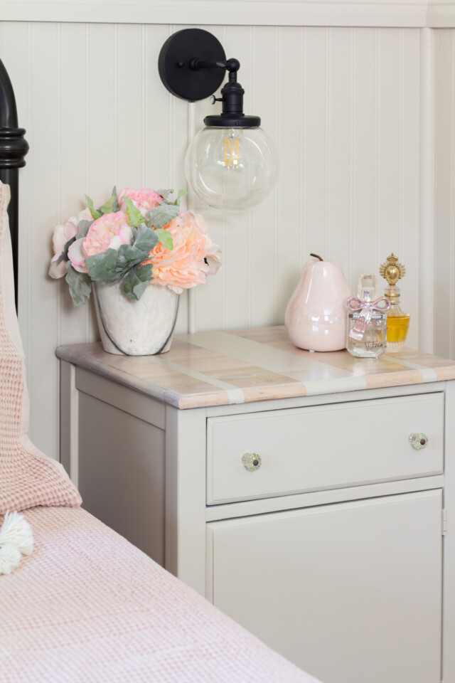 Staged bedside table, beside a bed with a pink comforter, topped with a flower bouquet and glass figurines