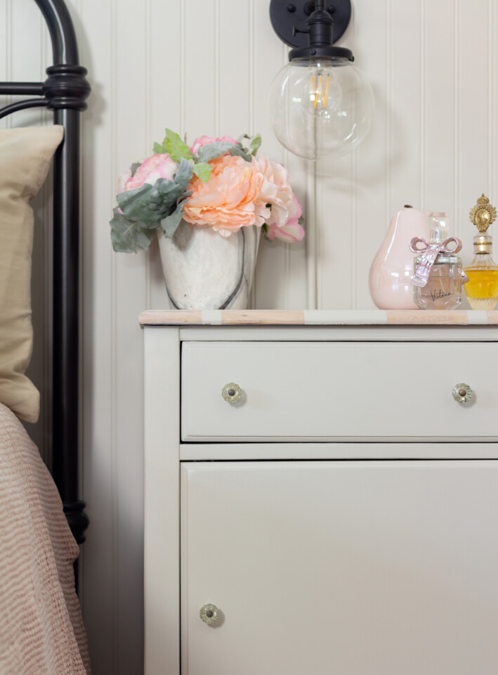 Staged bedside table, beside a bed with a pink comforter, topped with a flower bouquet and glass figurines