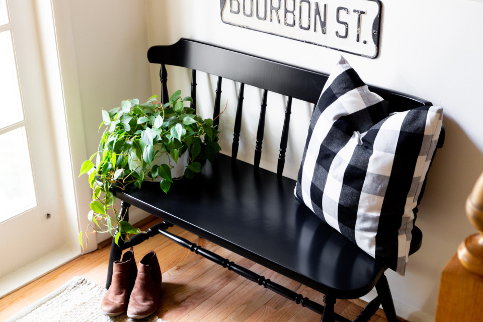 Staged entry way bench in black with black and white plaid pillow, boots and a plant
