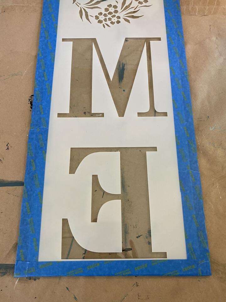 Close up taped home stencial, only the M and E are visible taped down with blue painters tape