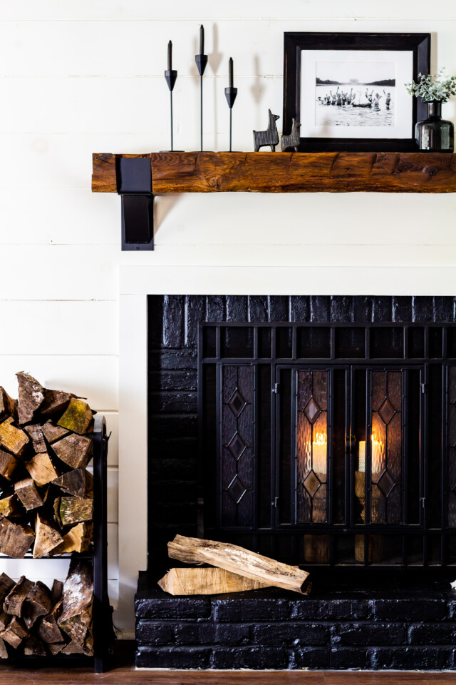 staged painted fireplace, black fireplace with black brick, stack of firewood, a throw blanket, wooden shelf above with a picture frame and other black modern decors
