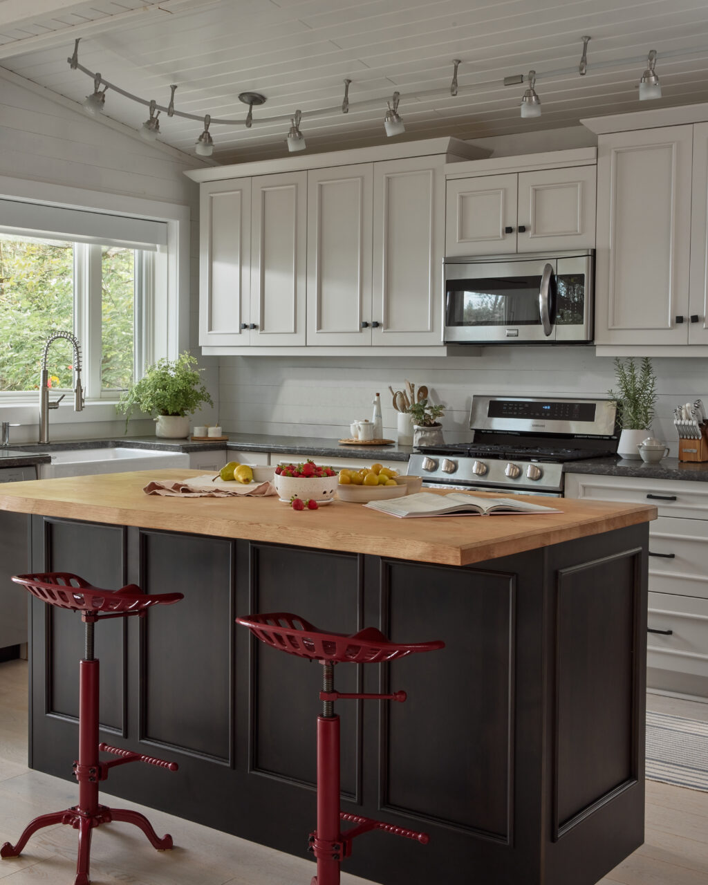 Kitchen with white cabinets, dark grey island and red stools. Staged with fresh fruit & a recipe book on the island.