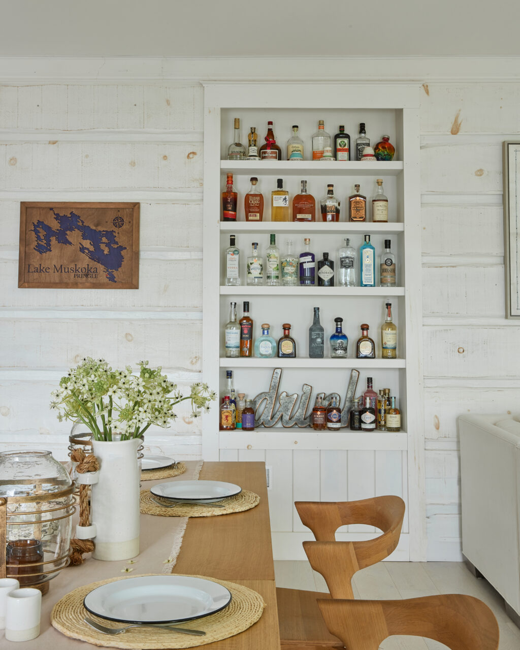 Wooden dining room table, set with beige placemats and white plates, in the back their is white shelving with a variety of alcohol bottles