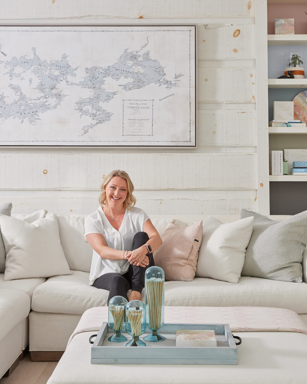 Cottage Makeover Series - Jennylyn sitting on white coach in front of white washed wood panel walls with a map of Muskoka Lakes on the wall