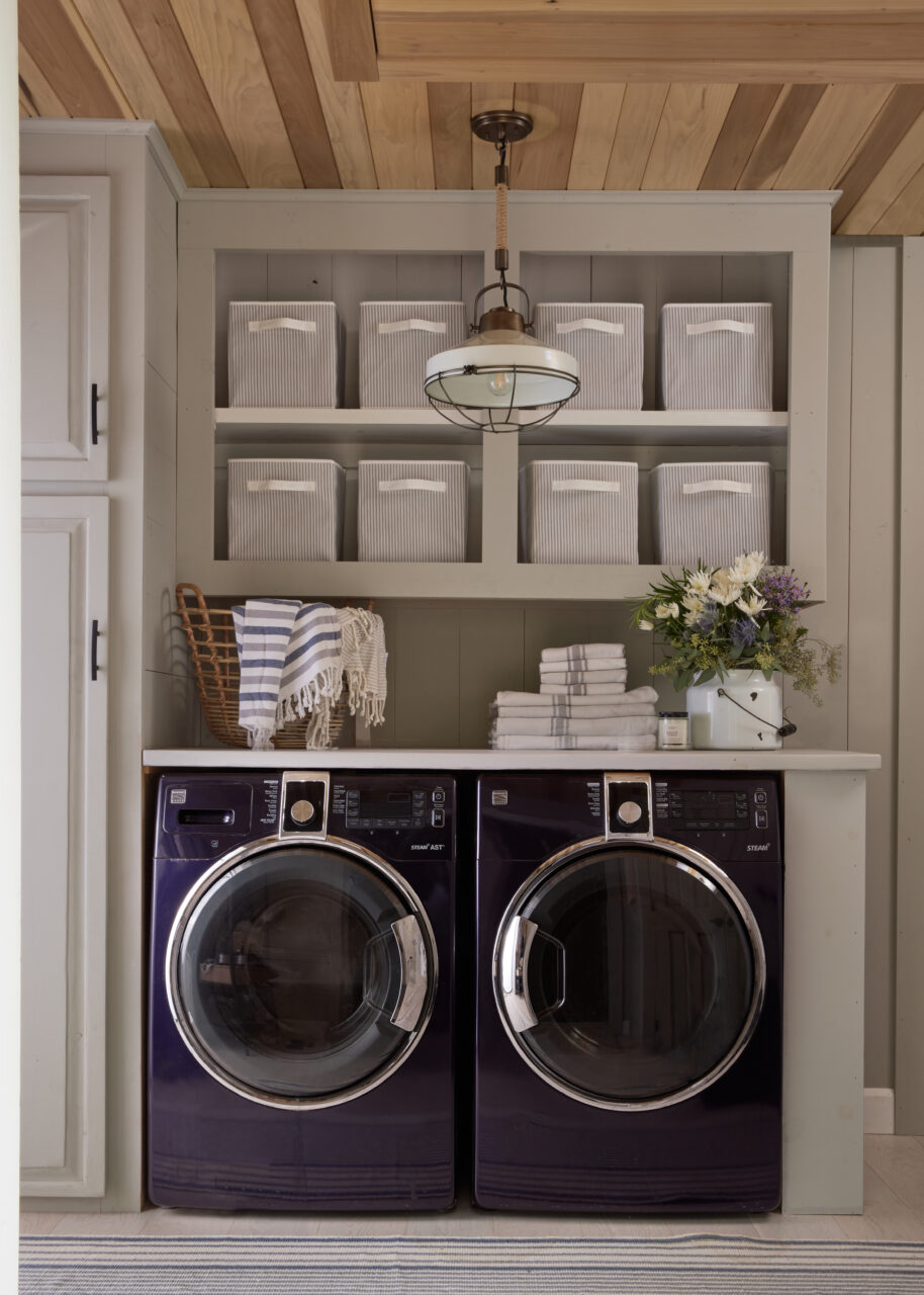 Laundry room with dark washer/dryer, light walls & cabinets and grey baskets on shelves