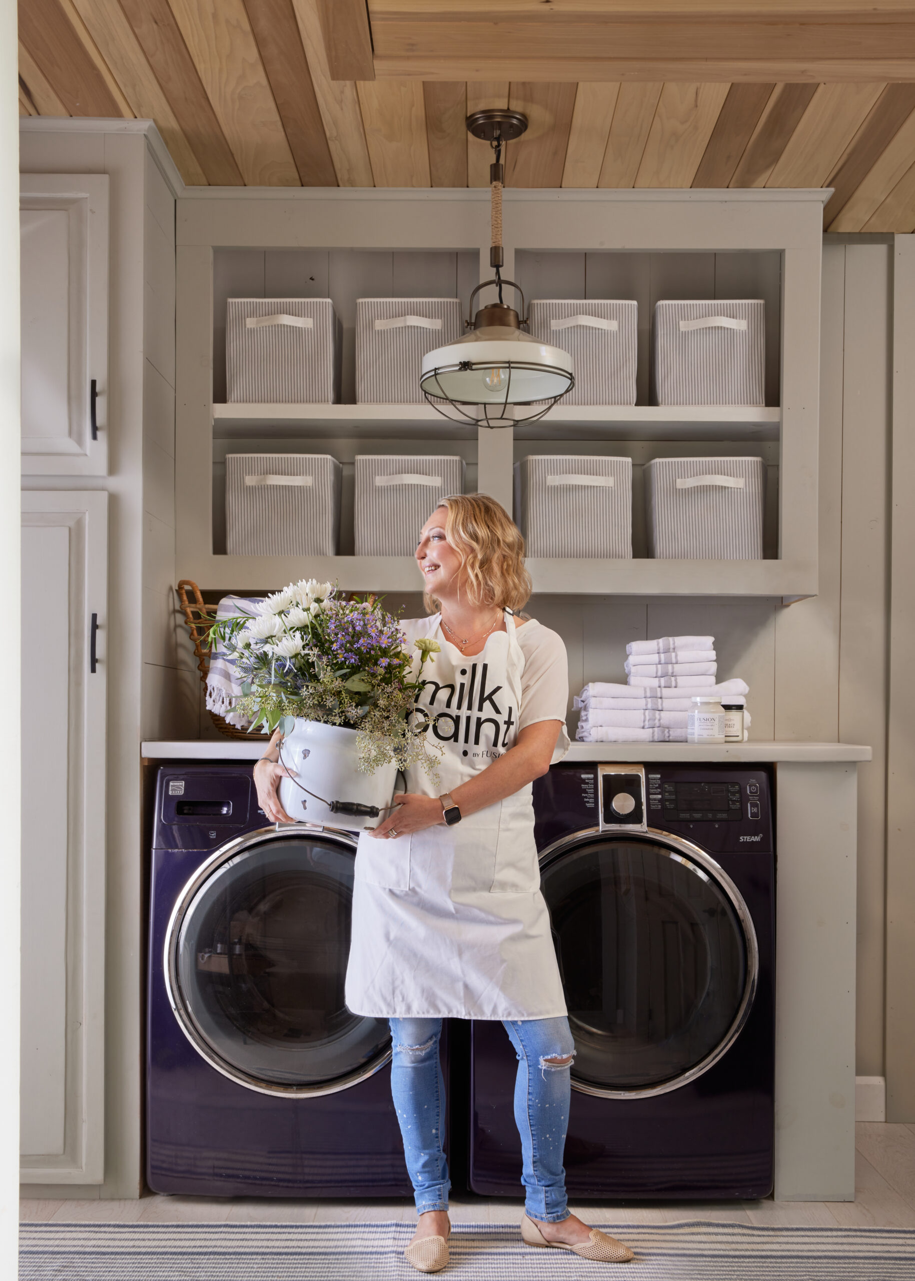 Jennylyn holding a bouquet of flowers, wearing a milk paint apron & standing in front of dark washer/dryer with light coloured walls/cabinets & grey baskets
