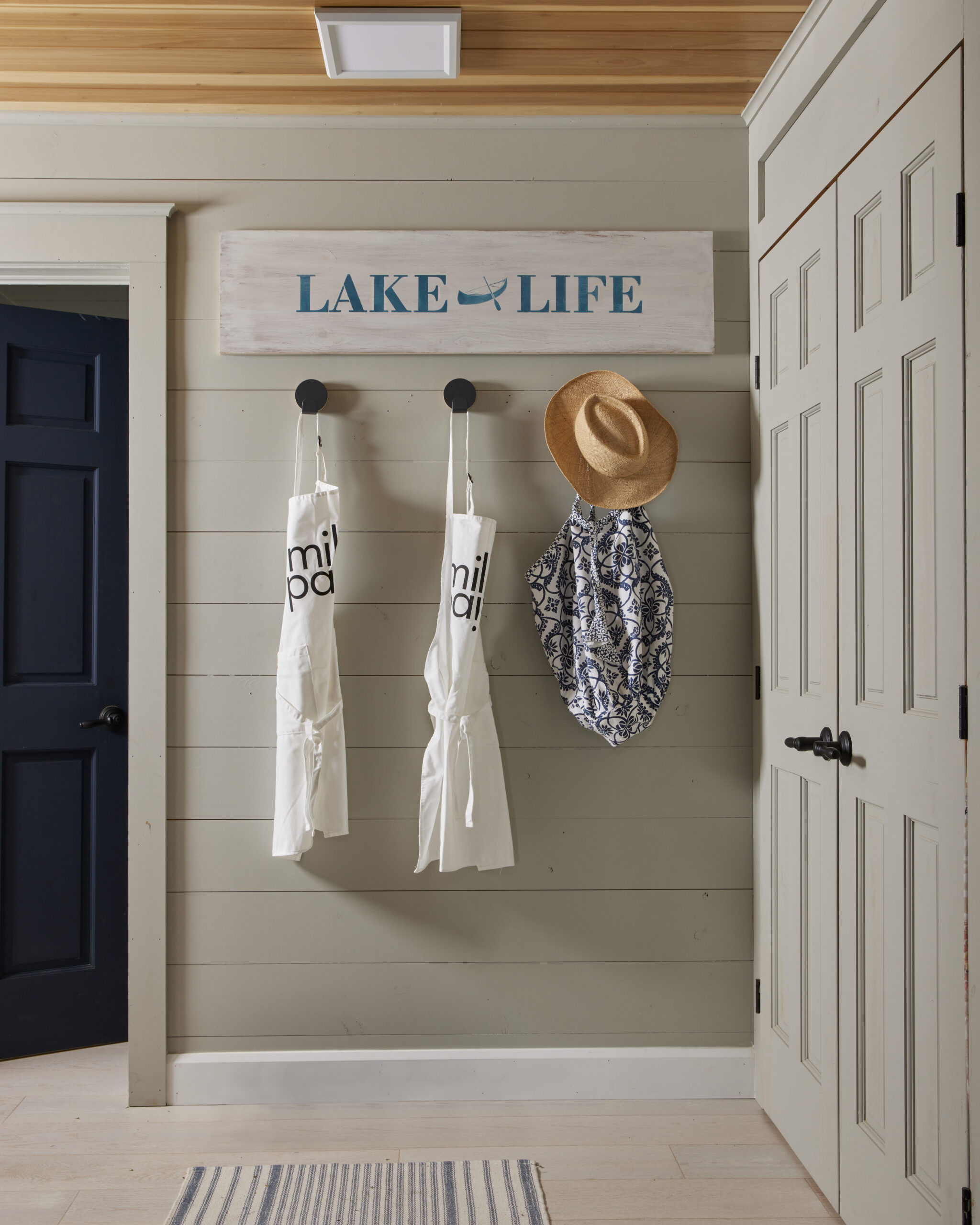 Entrance way to cottage with a sign that reads "lake life", hooks below the sign have a sun hat & coverup as well as two milk paint aprons hanging
